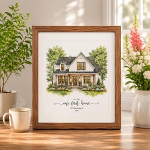 May include: A framed watercolor print of a two-story house with white siding and a dark roof, surrounded by green trees and flowers. Below the house, the text reads: Est. 2026, our first home, 197 Bainbridge St, 2026. The print is in a brown frame.