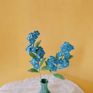 May include: A handmade bouquet of blue crochet forget-me-nots with yellow centres and green leaves, in a teal ceramic vase. The vase rests on a white lace cloth against a yellow backdrop. A charming, handmade floral arrangement.