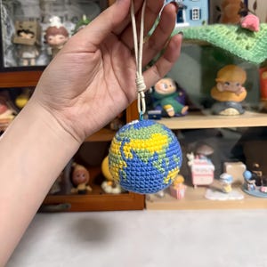May include: A crocheted globe keychain featuring blue oceans and yellow and green continents. The handmade Earth-shaped ornament hangs from a cream-colored cord. The globe is held up by a hand.