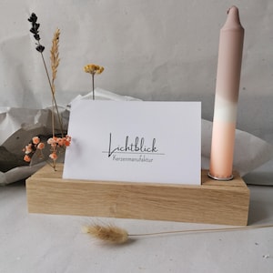 Könnte beinhalten: Ein Holzblock hält eine weiße Karte mit dem Text "Lichtblick Kerzenmanufaktur" und eine hohe Ombre-Kerze in Rosa- und Weißtönen. Getrocknete Blumen und Gräser verleihen der Präsentation eine natürliche Note.