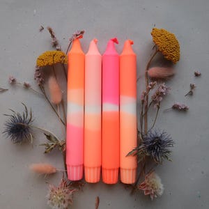 Puede incluir: Cinco velas de color neón con un efecto degradado, que transita del naranja al rosa y al blanco. Las velas están dispuestas con flores secas y follaje sobre una superficie gris neutra, creando una exhibición vibrante y artística. Las velas son un artículo decorativo.