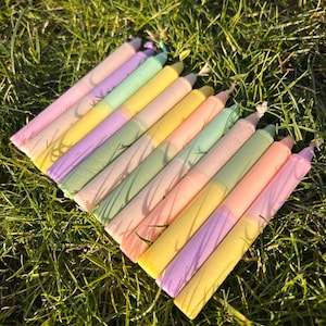 Könnte beinhalten: Eine Sammlung pastellfarbener Kerzen, die auf einer grünen Grasfläche angeordnet sind. Die Kerzen sind in den Farbtönen Rosa, Gelb, Grün und Lila gehalten. Die Kerzen sind zylindrisch und scheinen handgefertigt zu sein. Die Gesamtästhetik ist weich und skurril.