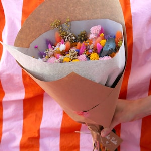 Könnte beinhalten: Ein farbenfroher Strauß Trockenblumen, verpackt in braunem Papier. Der Strauß enthält Blumen in Rosa, Orange, Gelb und Lila. Er ist mit Bindfaden und einem kleinen Anhänger gebunden.