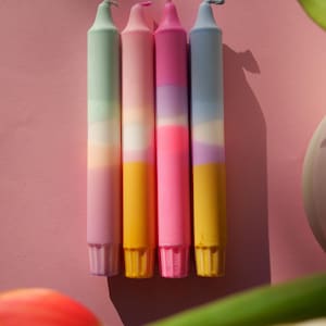 Puede incluir: Cuatro velas cónicas coloridas dispuestas sobre una superficie rosa. Cada vela presenta un degradado de colores, incluyendo verde, rosa, morado y amarillo. Las velas no están encendidas y tienen una forma cónica clásica. La estética general es brillante y alegre.