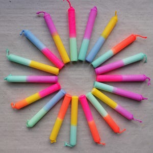 Könnte beinhalten: Kreisförmige Anordnung farbenfroher Kerzen. Jede Kerze hat ein Farbverlaufsdesign, das von leuchtenden Farben wie Pink, Orange und Gelb zu sanfteren Blau- und Grüntönen übergeht. Die Kerzen sind auf einer neutralen Oberfläche angeordnet.