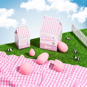 May include: Pink Cutie Blenders makeup sponges and milk carton-shaped packaging with the text "Cutie Blenders" and a cow illustration. The scene includes a pink and white checkered picnic blanket, toy cows, and a white picket fence on a green lawn.
