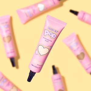 Puede incluir: Tubos rosas de base de sombra de ojos con tapas negras dispersos sobre un fondo amarillo. Los tubos tienen las palabras "MAKE EM POP" y "EYESHADOW BASE" impresas, junto con un diseño de corazón. Cada tubo contiene 15 ml de producto.