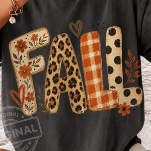 May include: A charcoal gray t-shirt featuring the word FALL in large letters. The F has orange flowers, the A has a leopard print, the L has an orange plaid pattern, and the second L has black polka dots. A small heart and leaf are included.