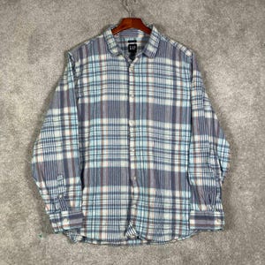 Puede incluir: Una camisa de manga larga con botones de la marca Gap que presenta un patrón de cuadros en tonos azul, blanco y naranja. La camisa tiene un cuello estándar, un cierre frontal completo de botones y puños abotonados sobre una percha.