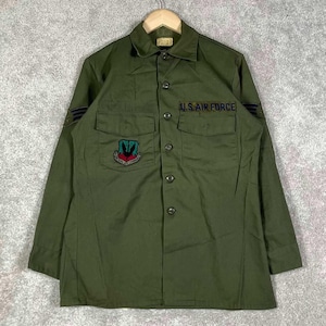 Può includere: Una camicia a maniche lunghe abbottonata color verde oliva con una toppa U.S. AIR FORCE sopra la tasca destra del petto e una toppa TACTICAL AIR COMMAND sulla tasca sinistra. La camicia presenta toppe a gallone scure su entrambe le maniche.
