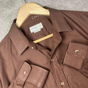May include: A solid brown long-sleeve button-up shirt with a pointed collar and cuffs. The shirt features a white label on the inside neck with the text Marcia Freer, Fine Apparel, Little Rock, Ark, 100% Pure Cotton. The word Gray is handwritten.