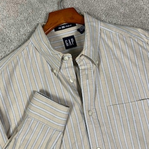 Könnte beinhalten: Ein langärmeliges Button-Down-Hemd in Beige mit vertikalen weißen und blauen Streifen. Das Hemd verfügt über einen Button-Down-Kragen, eine Brusttasche und ein dunkelblaues Etikett am Kragen mit der Aufschrift GAP und The Big Oxford.