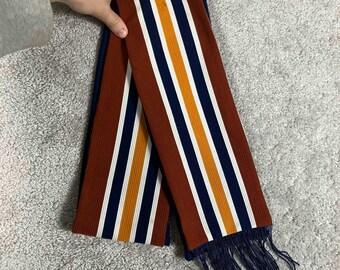 May include: A long, rectangular knit scarf featuring a repeating vertical striped pattern in rust orange, white, navy blue, and mustard yellow. The scarf has a ribbed texture throughout and is finished with navy blue fringe at the bottom edge.