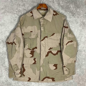 Könnte beinhalten: Beige-braune Tarnjacke im Wüstenmuster mit vier Fronttaschen, geknöpften Manschetten und einem Kragen. Die Jacke hängt an einem Holzkleiderbügel vor einem grauen Hintergrund. Das Tarnmuster ist im Wüstenstil gehalten.