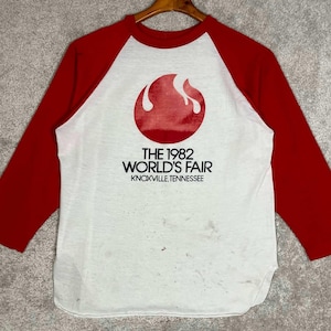 May include: A white baseball-style t-shirt with red three-quarter length sleeves. The front features a red circular flame graphic above the black text THE 1982 WORLD'S FAIR KNOXVILLE, TENNESSEE. The white fabric shows faint scattered dark spots.