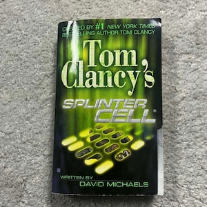 Op de afbeelding: Een paperback boek getiteld "Tom Clancy's Splinter Cell" door David Michaels. De cover heeft een groen en zwart ontwerp met de titel in grote, vette letters. Het boek is van #1 New York Times bestsellerauteur Tom Clancy.