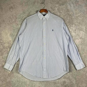 May include: A long-sleeved button-down shirt with vertical blue and white stripes. The shirt features a button-down collar and a small navy blue logo on the chest. The shirt is hanging on a wooden hanger.