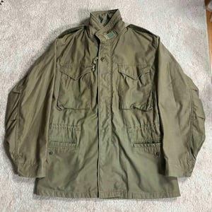 May include: Olive green military-style jacket with a high collar and multiple pockets. The jacket has a button and zipper closure, epaulets, and a drawstring waist. The fabric appears to be a durable, medium-weight material.