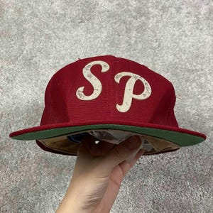 Puede incluir: Una gorra de béisbol granate con las letras S y P en fieltro blanco roto desgastado en la parte delantera. La gorra tiene una corona estructurada, una visera plana con el reverso verde oscuro y un forro interior de color tostado.