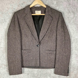 Puede incluir: Blazer marrón de espiga con solapas de muesca, dos bolsillos delanteros y cierre de un solo botón. El blazer está colgado de una percha de madera. El forro es negro y la tela tiene un aspecto texturizado.