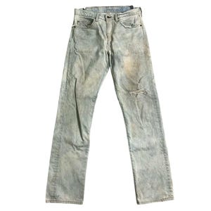 Puede incluir: Vaqueros de mezclilla azul claro con un aspecto desgastado y descolorido. Los vaqueros tienen una rodilla rasgada y muestran signos de desgaste. Los vaqueros están colocados planos sobre un fondo blanco.