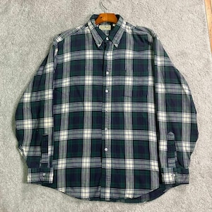 Puede incluir: Camisa a cuadros verdes, azul marino y blancos. La camisa de manga larga tiene cuello abotonado y bolsillo en el pecho. La camisa está colgada de una percha de madera.