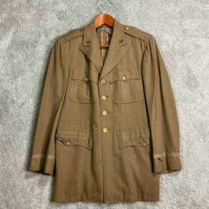 May include: A tan military jacket with gold buttons and four front pockets. The jacket has the letters "U.S." on the shoulders and is hanging on a wooden hanger. The jacket is a vintage style.