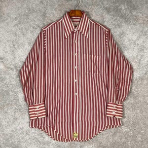 Op de afbeelding: Een shirt met lange mouwen met verticale rood-witte strepen. Het shirt heeft een puntkraag, een borstzak en manchetten met knopen. Het shirt hangt aan een houten kleerhanger.