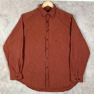 May include: A long-sleeve button-down shirt in a rust-orange and dark brown micro-check pattern. The shirt features a standard collar, a single chest pocket on the left side, a full-length front button placket, and buttoned cuffs on the sleeves.