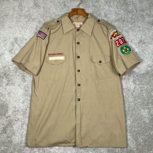 May include: Tan short-sleeve shirt with a button-down front, two chest pockets, and patches. The patches include an American flag, the words "BOY SCOUTS OF AMERICA", and a patch with the number "201".