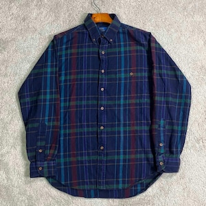Può includere: Una camicia abbottonata a maniche lunghe con un motivo a quadri. La camicia presenta una base blu navy con strisce verticali e orizzontali turchese, bordeaux e marroni. Ha un colletto button-down e una tasca sul petto.