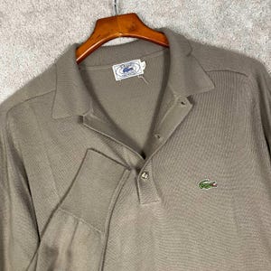 May include: A long-sleeve taupe polo shirt featuring a ribbed collar, a buttoned placket, and a small embroidered green crocodile logo on the chest. The interior neck label displays the text IZOD LACOSTE with a size L indicator visible.