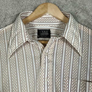 May include: A long-sleeve button-down shirt featuring a white base with vertical brown stripes and a repeating checkered pattern. The shirt has a pointed collar and a black rectangular label at the inner neckline with the white text XKE printed on it.