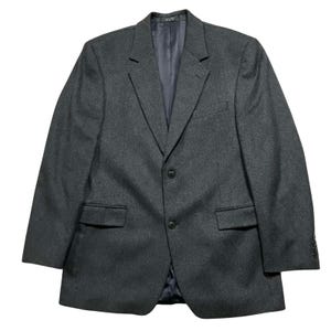 Könnte beinhalten: Ein anthrazitfarbener Blazer mit Zwei-Knopf-Verschluss, fallendem Revers und zwei Vordertaschen. Die Jacke hat ein dunkelblaues Futter und ist aus einem strukturierten Stoff gefertigt. Ein klassischer Stil.