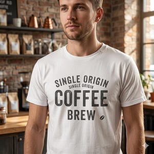 Puede incluir: Una camiseta blanca con las palabras "SINGLE ORIGIN COFFEE BREW" impresas en negro. La camiseta tiene cuello redondo y mangas cortas. Un gráfico de grano de café está debajo de la palabra "BREW". La persona está en una cafetería.