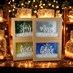 Puede incluir: Cuatro decoraciones navideñas enmarcadas con las palabras "joy", "Merry", "hope" y "believe" en escritura blanca. Cada marco tiene un fondo de color diferente y un diseño de copo de nieve. Los marcos son de madera clara.