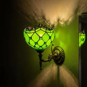 May include: A wall-mounted lamp featuring a bowl-shaped shade made of green stained glass panels arranged in a diamond grid pattern. The lamp has a dark bronze-toned metal base with ornate scrollwork and a circular mounting plate attached to the wall.
