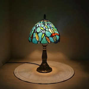 May include: A Tiffany-style table lamp with a stained glass shade. The shade features a dragonfly design in shades of blue, green, and orange. The base is a dark bronze colour. The lamp is on a round, off-white surface.