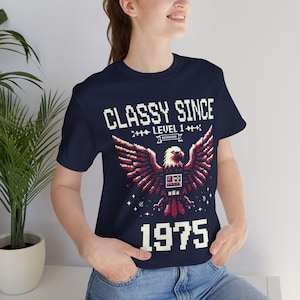 Könnte beinhalten: Marineblaues T-Shirt mit einem pixeligen Adlerdesign in Rot und Weiß. Das Shirt zeigt den Text "CLASSY SINCE LEVEL 1" über dem Adler und das Jahr "1975" darunter. Ein lässiges Grafik-T-Shirt im Retro-Stil.