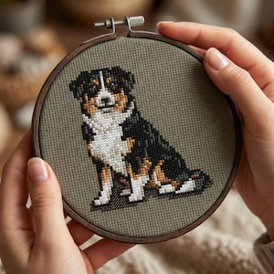 Op de afbeelding: Kruissteekborduurwerk van een hond in een houten hoepel. De hond is zwart, bruin en wit, met een gedetailleerd ontwerp. De achtergrond is een gedempte groene stof. De hoepel wordt door handen vastgehouden.