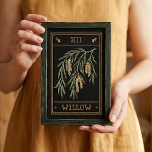 Op de afbeelding: Een ingelijste kruissteek met een wilgentak met groene bladeren en gele katjes tegen een zwarte achtergrond. De tekst "XII" staat bovenaan en "WILLOW" onderaan. De lijst is donkergroen.