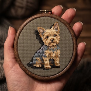 Può includere: Un telaio da ricamo circolare in legno contenente un motivo a punto croce di un piccolo cane con pelo beige e grigio. Il cane è raffigurato in posizione seduta su uno sfondo di tessuto verde oliva, incorniciato da un cerchio in legno scuro.