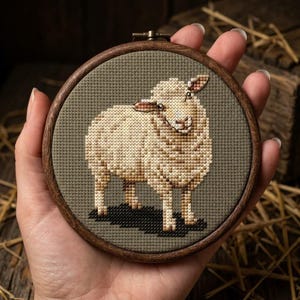 May include: A circular wooden embroidery hoop containing a cross-stitch pattern of a cream-coloured sheep with brown shading. The sheep is centred on a dark olive green fabric background, featuring a black shadow cast beneath its four legs.