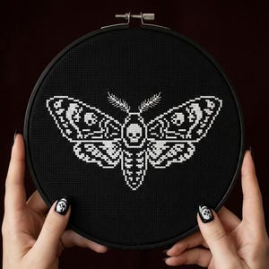 May include: A black cross-stitch embroidery hoop featuring a white moth design with skull details. The moth is centered on the black fabric, and the hoop is held by hands with black nail polish and skull nail art.