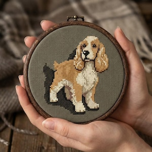Könnte beinhalten: Kreuzstich-Stickerei eines Cocker Spaniel-Hundes in einem Holzreifen. Der Hund ist in Braun-, Beige- und Weißtönen vor einem grauen Hintergrund dargestellt. Der Reifen wird von Händen gehalten, und die Gesamtästhetik ist rustikal und handgefertigt.