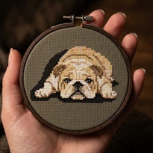 Pode incluir: Um bastidor de bordado em ponto cruz com um cão castanho claro e branco com uma sombra escura. O cão está deitado sobre um fundo de tecido verde. O bastidor de madeira é segurado em uma mão.