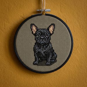 Può includere: Ricamo a punto croce di un bulldog francese nero in un cerchio di legno rotondo. Il cane è centrato su uno sfondo di tessuto beige. Il cerchio è sospeso da uno spago di spago naturale contro un muro giallo senape.