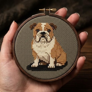 Pode incluir: Um bastidor de bordado circular de madeira contendo um padrão de ponto cruz de um bulldog castanho e branco. O cão está representado numa posição sentada sobre um fundo de tecido taupe sólido, com uma sombra escura sob as patas.