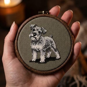 Op de afbeelding: Een ronde houten borduurring met een kruissteekontwerp van een staande terriër. De hond is geborduurd met grijze en witte draden op een olijfgroene stoffen achtergrond en toont een gedetailleerde weergave van de vacht.