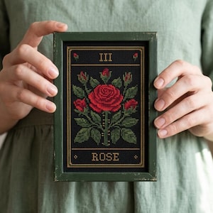 Puede incluir: Un bordado a punto de cruz enmarcado que presenta una rosa roja con hojas y capullos verdes sobre un fondo negro. El marco verde oscuro rodea el texto "III ROSE". La obra de arte está sostenida por una persona.